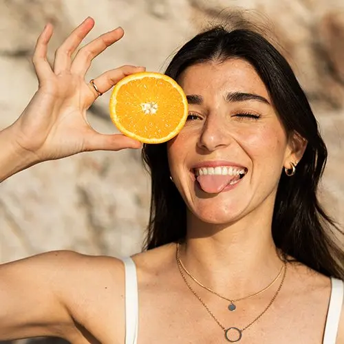 Mujer sonriente sosteniendo media naranja como símbolo de vitamina C y antioxidantes para el cuidado natural de la piel. 
