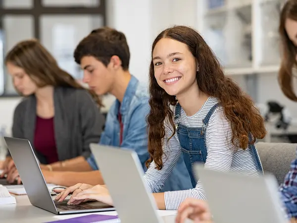 Vier Schüler sitzen am Schreibtisch und arbeiten am Laptop, Schülerin in der Mitte lächelt in die Kamera. 