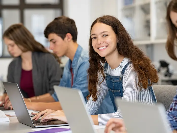 Vier Schüler sitzen am Schreibtisch und arbeiten am Laptop, Schülerin in der Mitte lächelt in die Kamera. 