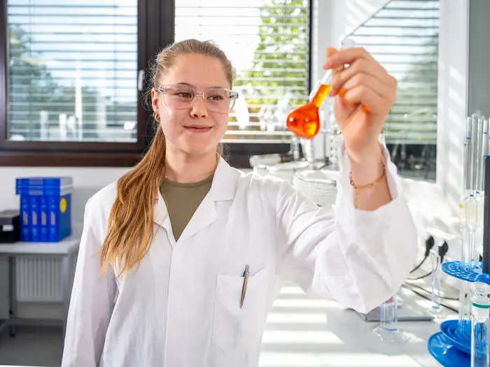 Eine Auszubildende Chemielaborantin in einem weißen Kittel und Schutzbrille steht im Labor und hält einen Kolben mit orangener Flüssigkeit in der Hand.
