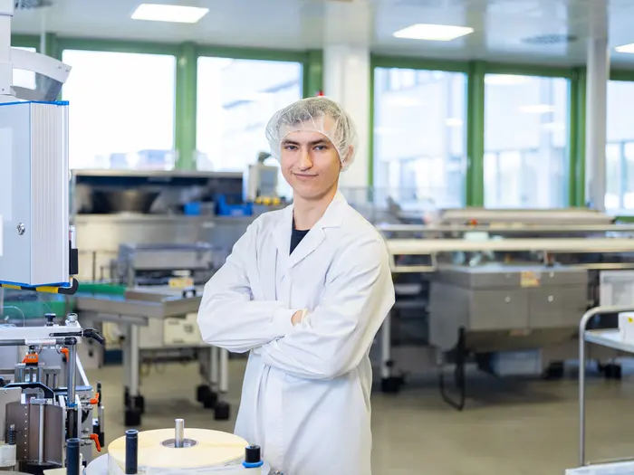 Ein Auszubildender Pharmakant in einem weißen Kittel und einer Haarhaube steht in einer Produktionsumgebung vor einer Maschine.