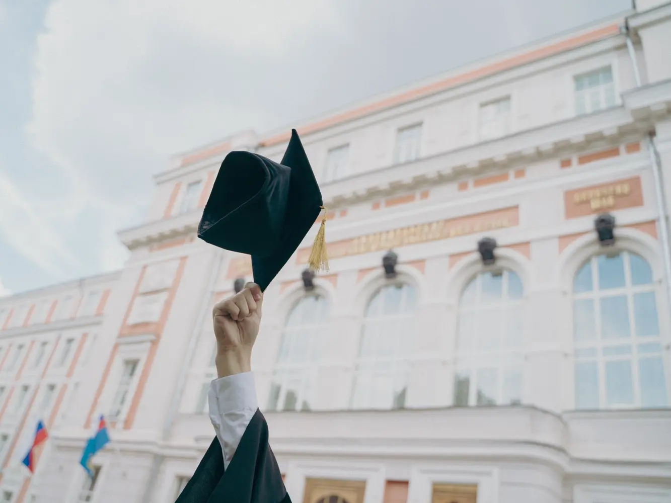Ein Absolvent steht vor einer Universität und hält seinen Absolventenhut in die Höhe. 