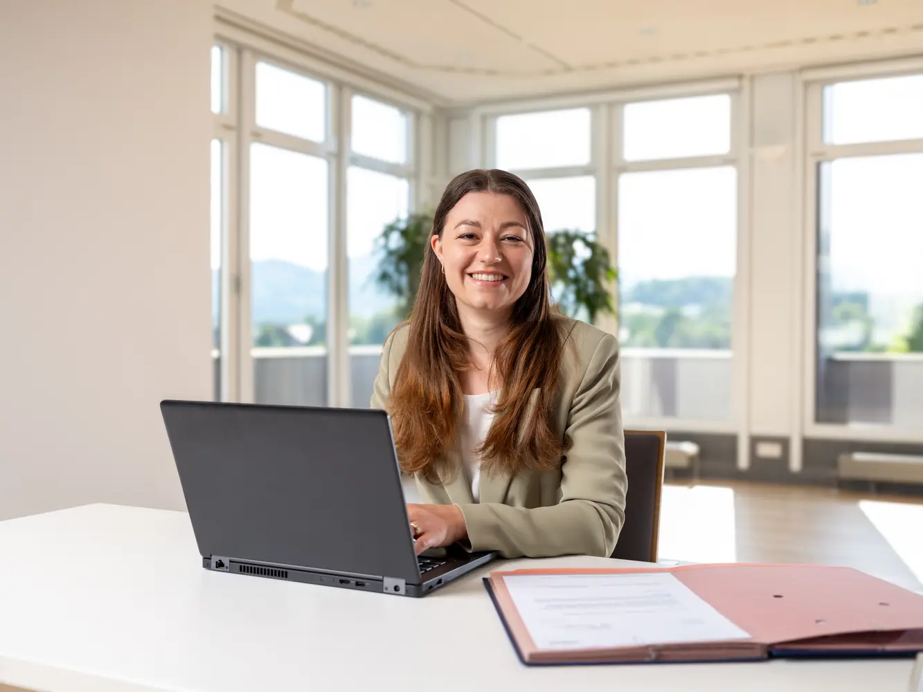 Kaufmännische Mitarbeiterin sitzt am Schreibtisch, tippt auf ihren Laptop und lächelt in die Kamera.