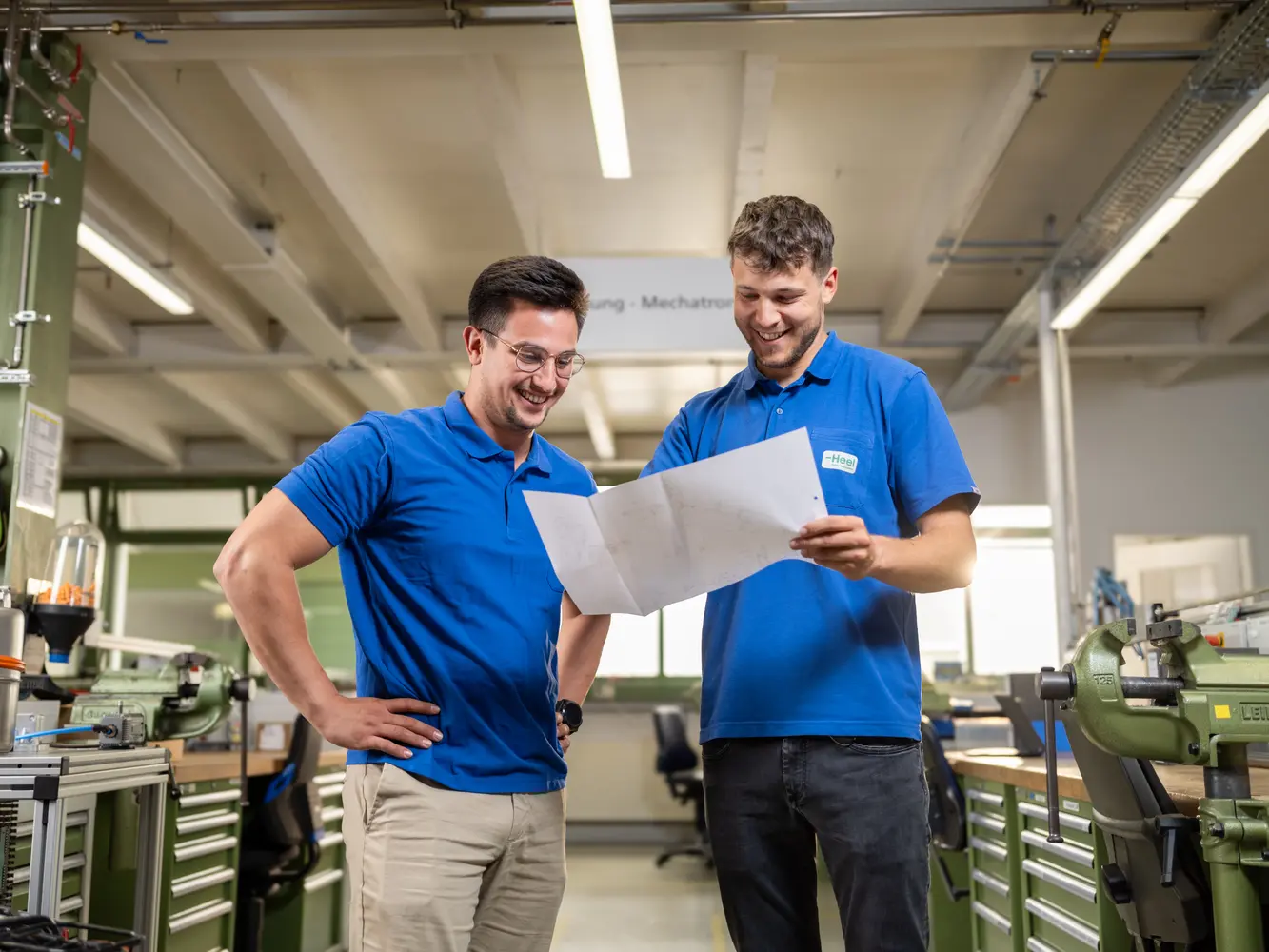 Zwei Mechatroniker im Bereich Technik stehen in Werkstatt und sehen sich einen Konstruktionsplan an, den einer der beiden in der Hand hält.