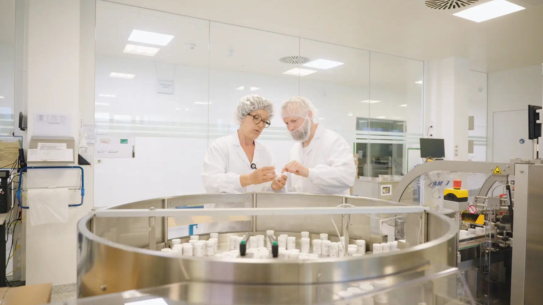 Zwei Pharmakanten in weißen Kitteln stehen hinter einer Produktionsanlage mit vielen kleinen Behältern darin und schauen sich eine Tablettenverpackung an.