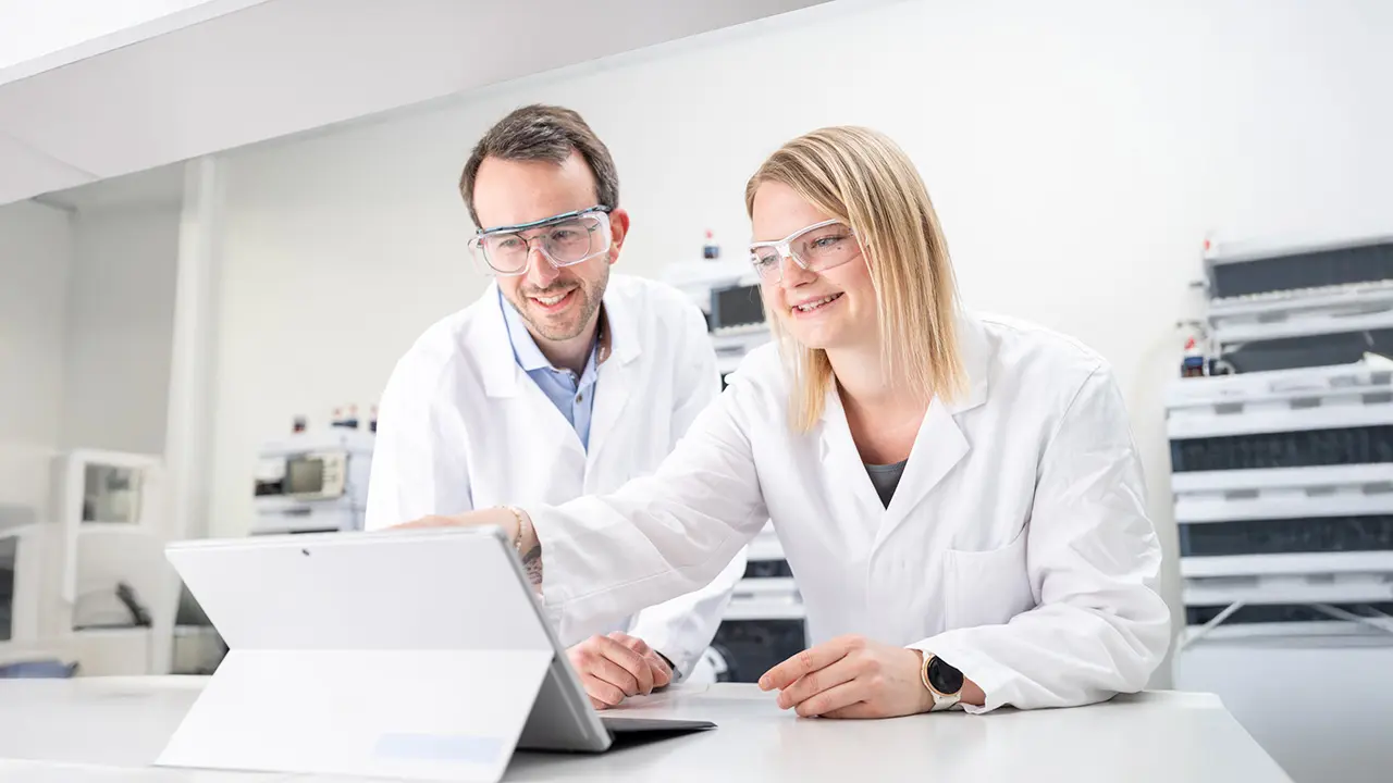 Zwei Ingenieure mit weißem Kittel und Schutzbrille stehen in Labor und schauen auf ein modernes Tablet.
